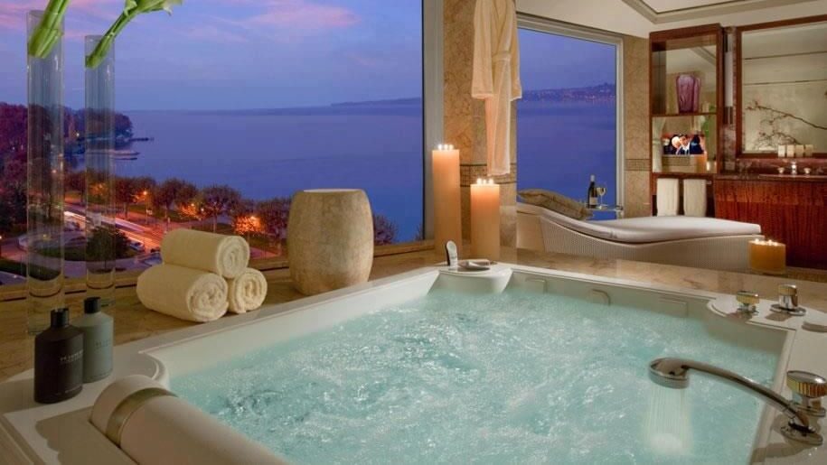 Luxury hotel bathroom with large Jacuzzi tub, candles, rolled towels, and panoramic waterfront view at dusk