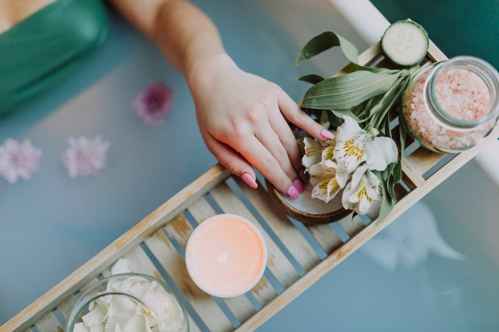 Hand resting on bath tray with candles, flowers, bath salts, and petals floating in a relaxing spa bath
