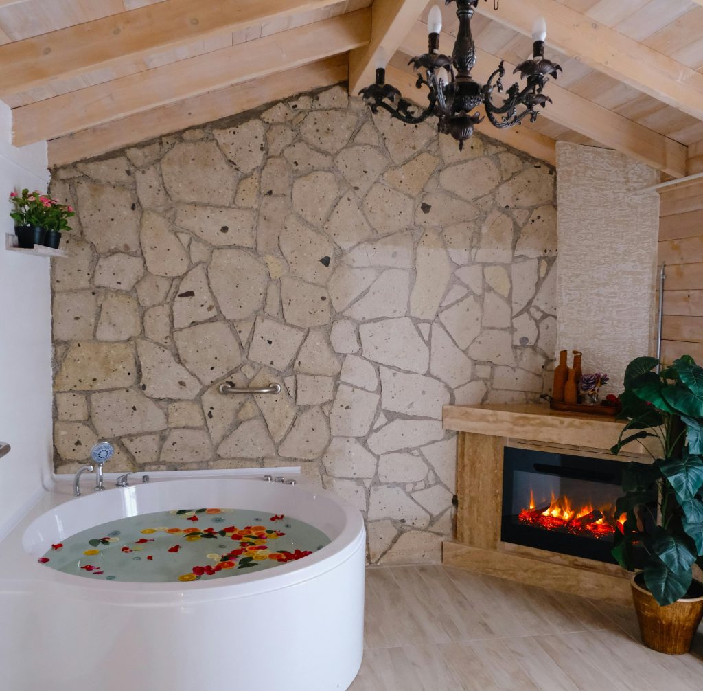 Luxury bathroom with round Jacuzzi tub, rose petals in water, stone walls, and fireplace ambiance