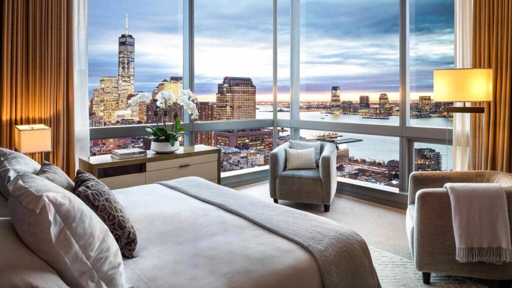 Hotel bedroom with large windows offering panoramic city views, armchairs, and soft evening light