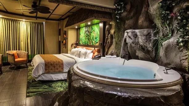 Themed hotel room with jungle decor, stone walls, and large in-room hot tub beside the bed