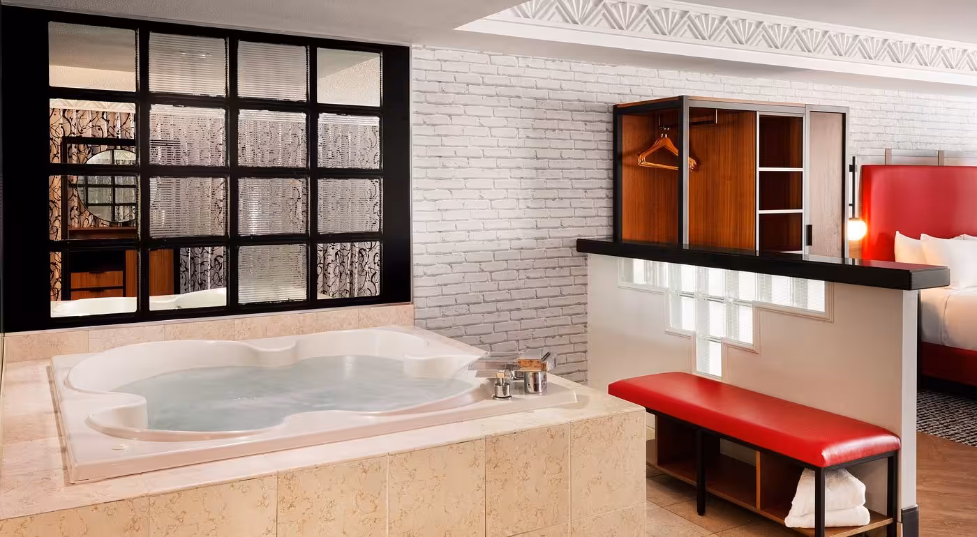 Modern hotel suite with in-room Jacuzzi tub, white brick wall, red accents, and open bedroom layout