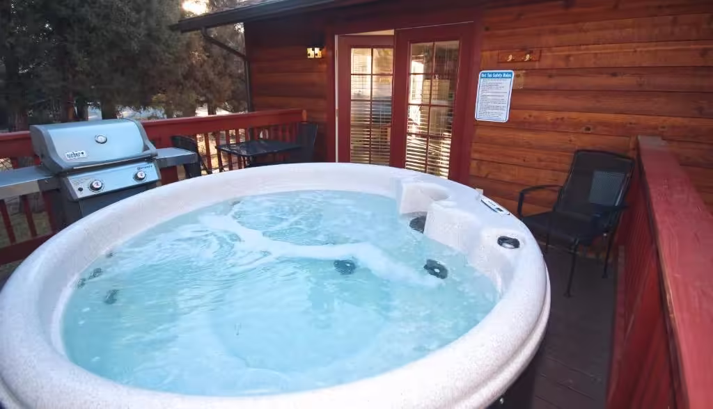Streamside on Fall River Outdoor hot tub on a private wooden deck outside a cabin, with grill, seating, and glass doors behind